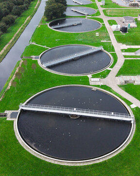 Aerial view of a wastewater treatment plant featuring large circular sedimentation tanks with metal bridges surrounded by green grass and a canal in Warmenhuizen, North Holland, Netherlands.