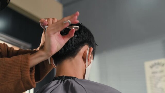 A barber cuts a seated customer&rsquo;s dark hair with scissors during a haircut. The barber holds a comb near the head while the customer wears a face mask and protective cape inside the salon.