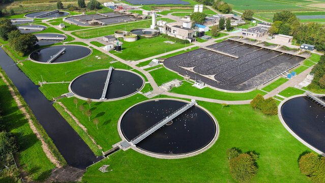 Aerial view of a wastewater treatment plant with circular sedimentation tanks and rectangular aeration basins surrounded by green lawns in Warmenhuizen, North Holland, Netherlands.
