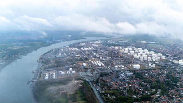 Aerial view of a large industrial oil refinery complex with numerous storage tanks and processing facilities situated along a riverbank near residential areas in Cilacap, Central Java, Indonesia.