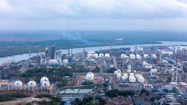 Aerial view of an industrial oil refinery complex featuring large storage tanks, flare stacks, and processing units situated near a river in Cilacap, Central Java, Indonesia.