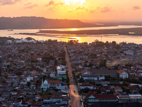 Aerial view of the coastal town of Cilacap at sunset, featuring dense residential rooftops, a prominent mosque, and a main road leading towards the bay in Cilacap, Central Java, Indonesia.