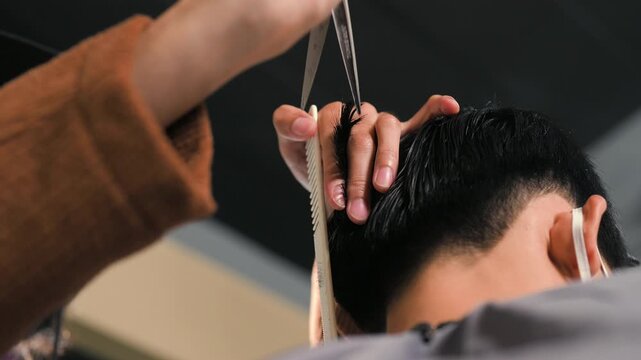 A barber cuts a seated customer&rsquo;s dark hair with scissors during a haircut. The barber holds a comb near the head while the customer wears a face mask and protective cape inside the salon.