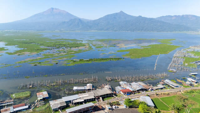 Aerial view of Lake Rawa Pening featuring water hyacinth patches, fish farms, and buildings along the shore with mountains in the background in Waduk Rawapening, Central Java, Indonesia.