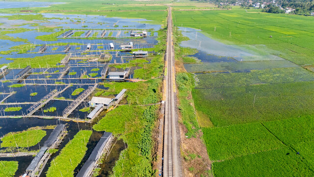 Aerial view of Lake Rawa Pening with a railway track dividing the water and lush green rice fields in Waduk Rawapening, Central Java, Indonesia.