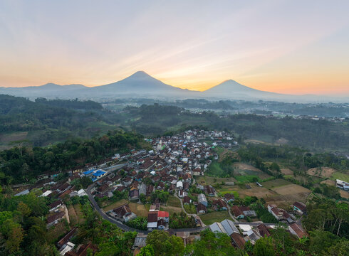 aerial view of a traditional Indonesian village nestled below Mount Sindoro and Sumbing during a beautiful golden misty sunrise in Wonosobo, Central Java.