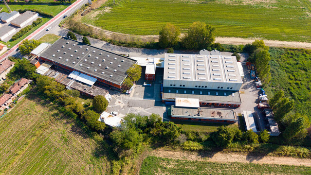 Aerial view of an industrial paint production facility with large warehouse buildings, skylights, and a paved yard surrounded by agricultural fields in Milan, Lombardy, Italy.