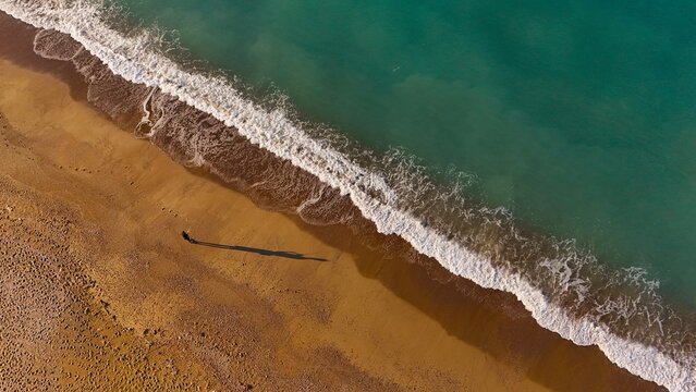 Aerial view of Manavgat Mendirek beach with turquoise sea waves washing onto the golden sand and a person walking casting a long shadow in Manavgat, Antalya, Turkiye.