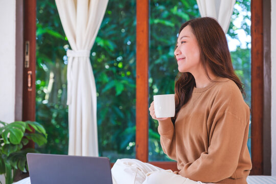 Portrait image of a woman working on laptop computer and drinking coffee while sitting on a bed at home
