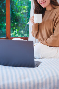 Vertical closeup image of a woman working on laptop computer and drinking coffee while sitting on a bed at home