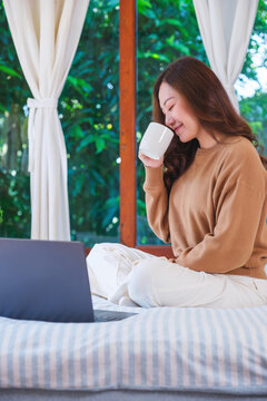Vertical portrait image of a woman working on laptop computer and drinking coffee while sitting on a bed at home