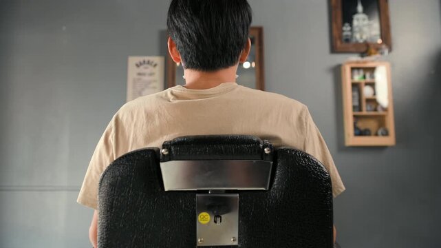 An empty barber chair sits inside a barbershop with black upholstery and a metal armrest. The barbershop interior shows the barber chair near a mirror, cabinet, bright window, and tiled floor.