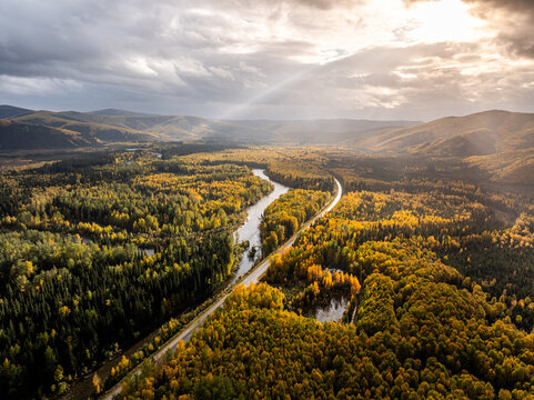 Aerial view of the Chena River winding through a dense forest with vibrant autumn foliage, under a dramatic sky with sunbeams in Chena, Alaska, United States.