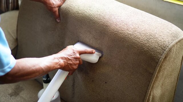 Close-up of person using handheld vacuum cleaner to clean textured fabric sofa upholstery, showing hands guiding device over worn surface with visible dirt spots and cleaning action.