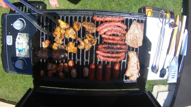 Person grilling assorted meat and sausages on outdoor barbecue grill at home.