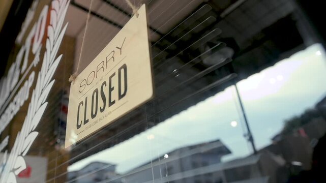 A closed sign hangs on the glass door of a barbershop. The barbershop entrance shows a hanging placard, storefront window, reflected light, and dark lettering against the clear glass.