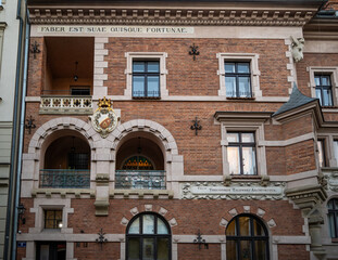 Obraz premium The detailed facade of a historical brick building showcases large arched openings, a balcony, and a prominent golden crest by architect Theodor Talowski in Krakow, Poland.