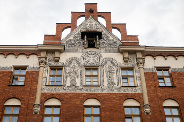 An old brick building features intricate white religious frescoes, arched windows, and decorative gables under a cloudy sky in Krakow, Poland. © Danica Chang