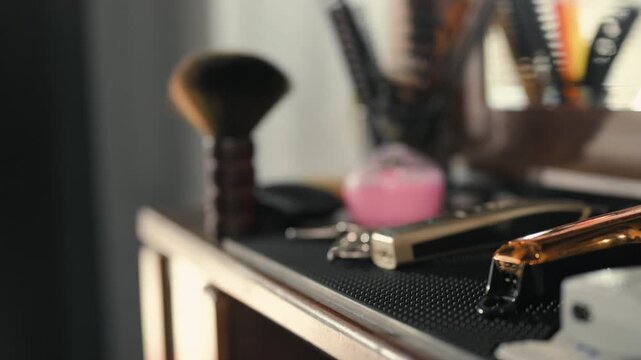 Grooming tools and electric clippers rest on a dark countertop inside a barbershop. The grooming tools include metal blades and clipper bodies, with a brush and small containers behind them.