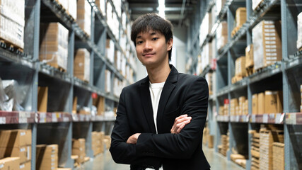 Fototapeta premium Smiling young Asian businessman standing with arms crossed in warehouse storage aisle. Successful e-commerce business owner and wholesale logistics concept.