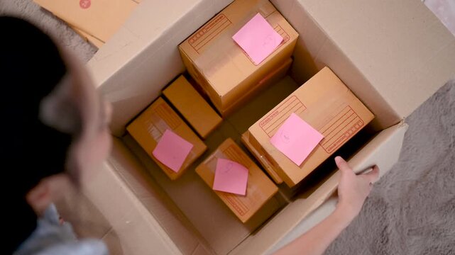 Cardboard boxes of different sizes form a parcel stack inside a home packing area. The cardboard boxes include open and closed brown cartons on the carpet beside a sofa and packing supplies.