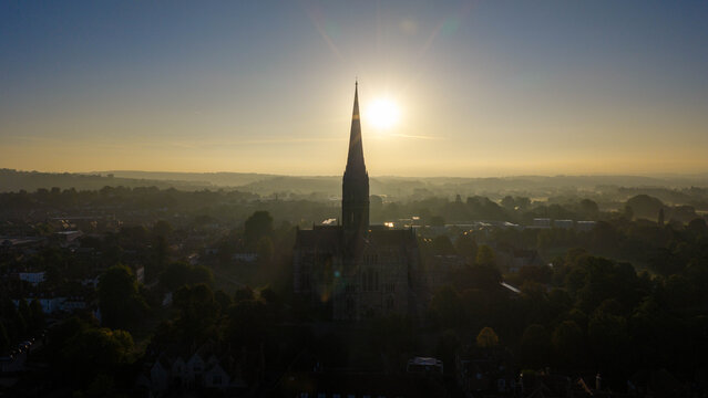 Aerial view of Salisbury Cathedral silhouetted against the rising sun with the historic city landscape in the background in Salisbury, England, United Kingdom.