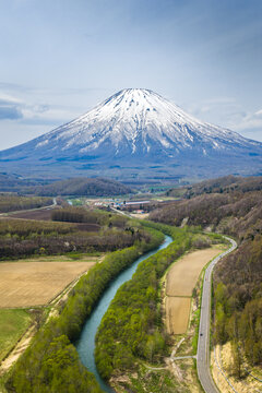 Aerial view of Mount Yotei with its snow-capped peak rising above the lush green forests, winding river, and agricultural fields near Niseko, Hokkaido, Japan.