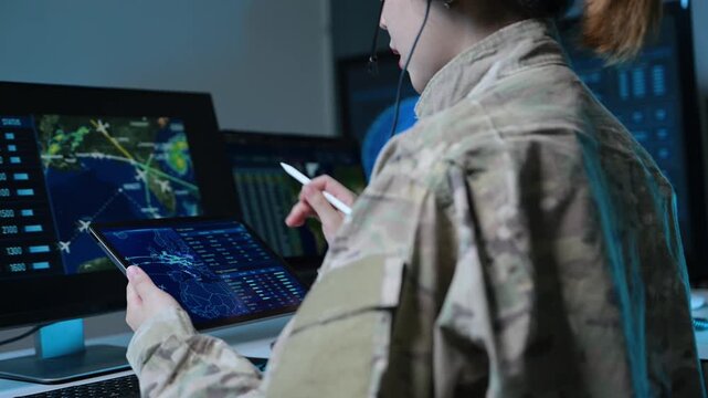 Asian female military officer in camouflage uniform analyzes radar maps on screens and writes notes in a high-tech tactical command center.