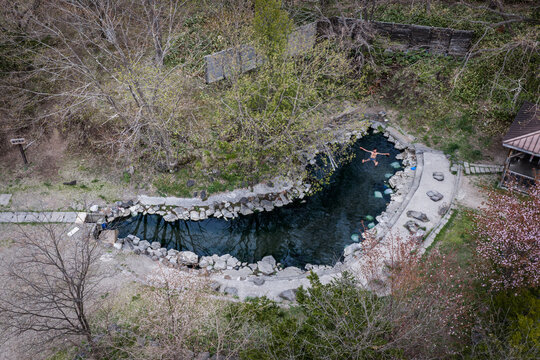 Aerial view of a person swimming in the natural stone-lined hot spring pool of Kotan Onsen at Lake Kussharo, surrounded by lush trees and rocky terrain in Teshikaga, Hokkaido, Japan.
