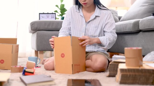 Cardboard boxes of different sizes form a parcel stack inside a home packing area. The cardboard boxes include open and closed brown cartons on the carpet beside a sofa and packing supplies.
