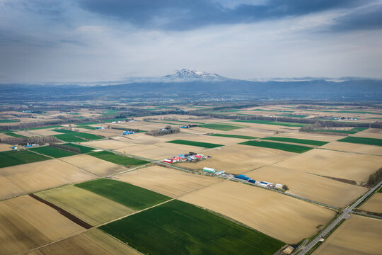 Aerial view of the agricultural landscape near Lake Kussharo, featuring patchwork fields, farm buildings, and distant snow-capped mountains under a cloudy sky in Teshikaga, Hokkaido, Japan.