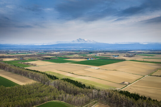 Aerial view of the agricultural landscape near Lake Kussharo with patchwork fields, forest belts, and distant snow-capped mountains under a cloudy sky, Teshikaga, Hokkaido, Japan.