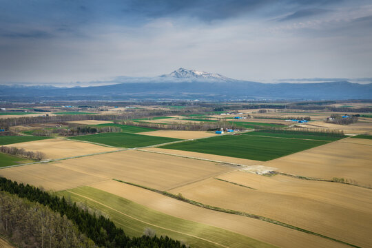 Aerial view of the agricultural landscape near Lake Kussharo with patchwork fields, distant snow-capped mountains, and a cloudy sky in Teshikaga, Hokkaido, Japan.