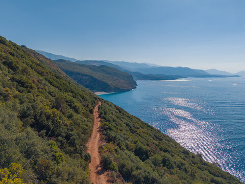 Aerial view of Gjipe Beach featuring a rugged dirt path winding along the lush green cliffside overlooking the sparkling blue Ionian Sea in Vlore County, Albania.