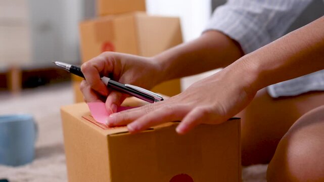 Hands place a shipping label on a brown parcel box while writing on a paper note. The hands work over the cardboard surface beside a pen and packing supplies in a close home workspace view.