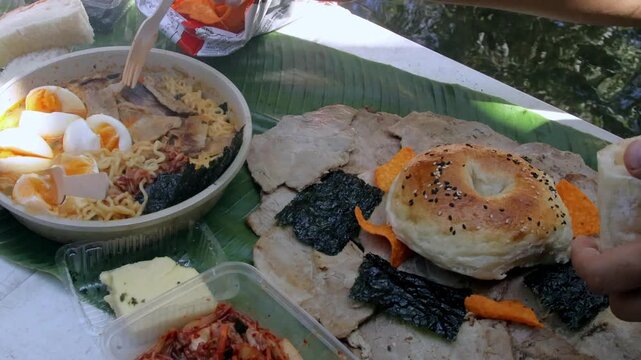 Overhead view stuffing bread with ramyeon noodles and egg, eating together in a korean picnic (sopung) by the river