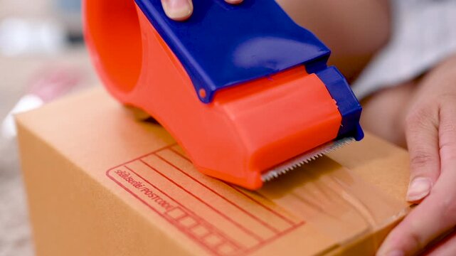 Hands press a tape dispenser against a parcel box to seal the cardboard surface. The hands guide clear packing tape across the box seam as the orange and blue tool moves over the brown carton.