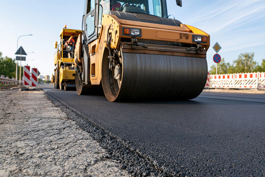 Yellow roller compacts fresh asphalt on new road surface. Worker monitors operation from truck cab. Roadside barriers guide traffic around construction zone