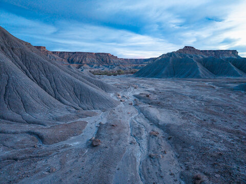 Aerial view of the rugged badlands of Caineville with eroded clay hills, dry riverbeds, and dramatic desert landscape under a soft blue sky in Caineville, Utah, United States.