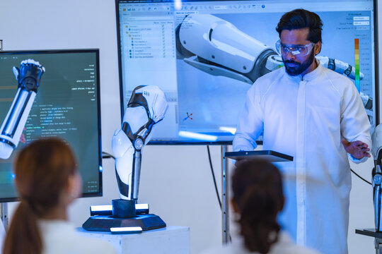 Bearded engineer in goggles presents a robotic arm stress heat map on a big screen, holding a tablet, during a problem-solving session, a next generation prosthetics concept