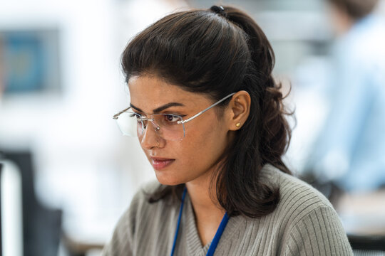 Close-up of Indian female engineer with glasses and ID lanyard focused on her work in a bright office, in an unposed working moment, an inclusive technology workplace scene