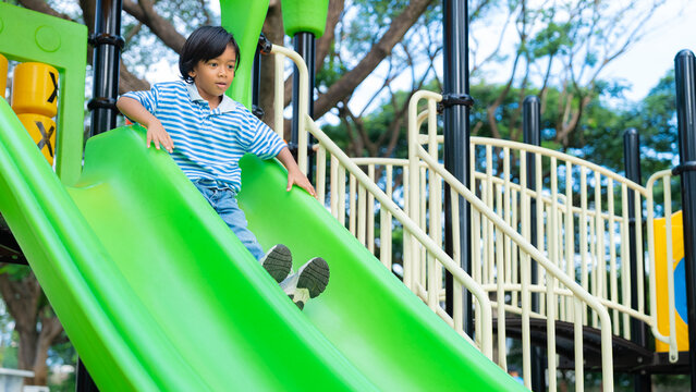 Active young Asian boy having fun sliding down a bright green slide at an outdoor playground. Healthy childhood and weekend recreation concept.