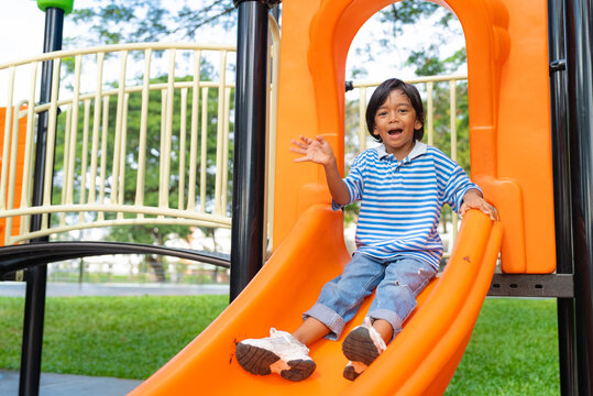 Happy young Asian boy waving hand and smiling while sliding down an orange slide at an outdoor playground. Active kids recreation and happy childhood concept.