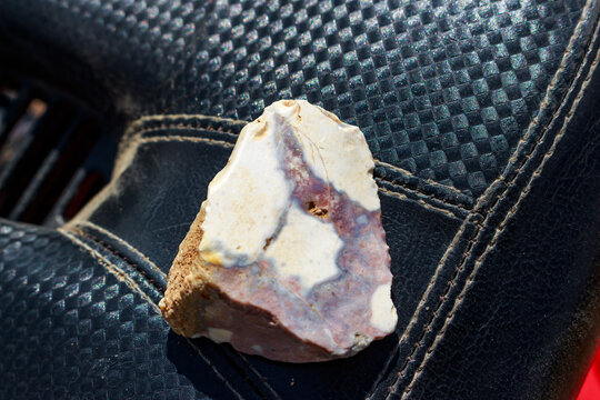 A weathered flint stone with a unique patina texture and cream-purple coloration rests on a black textured leather surface, capturing a raw geological detail in natural sunlight