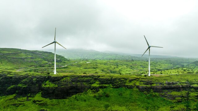 Aerial view of two wind turbines standing on a lush green cliffside under a cloudy sky in Igatpuri, Maharashtra, India.