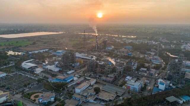 Aerial view of an industrial plant with complex machinery and smoking chimneys surrounded by fields under a warm sunset sky in Nagda, Madhya Pradesh, India.