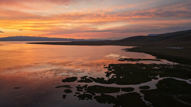 Aerial view of Song-Kul lake at sunset with vibrant orange and pink clouds reflecting on the calm water, surrounded by rolling hills and remote grasslands in Naryn Region, Kyrgyzstan.