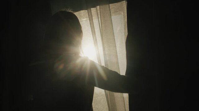 Silhouette of woman opening curtains in dark room as morning sun flares through window creating bright lens flare effect and warm atmosphere of new day beginning with cinematic slow motion lighting.