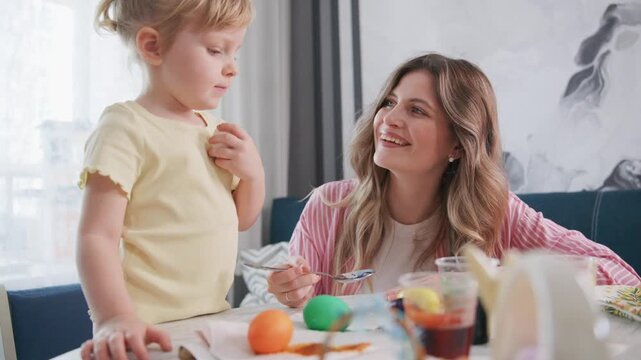 Mother smiles as she paints Easter eggs with her child. The child watches with wonder, holding her own brush. Bright colors fill the table around them