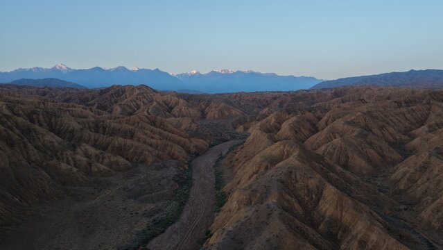 Aerial view of the rugged, arid canyons and rolling hills, with snow-capped mountains visible in the distance under a clear sky, Issyk-Kul Region, Kyrgyzstan.